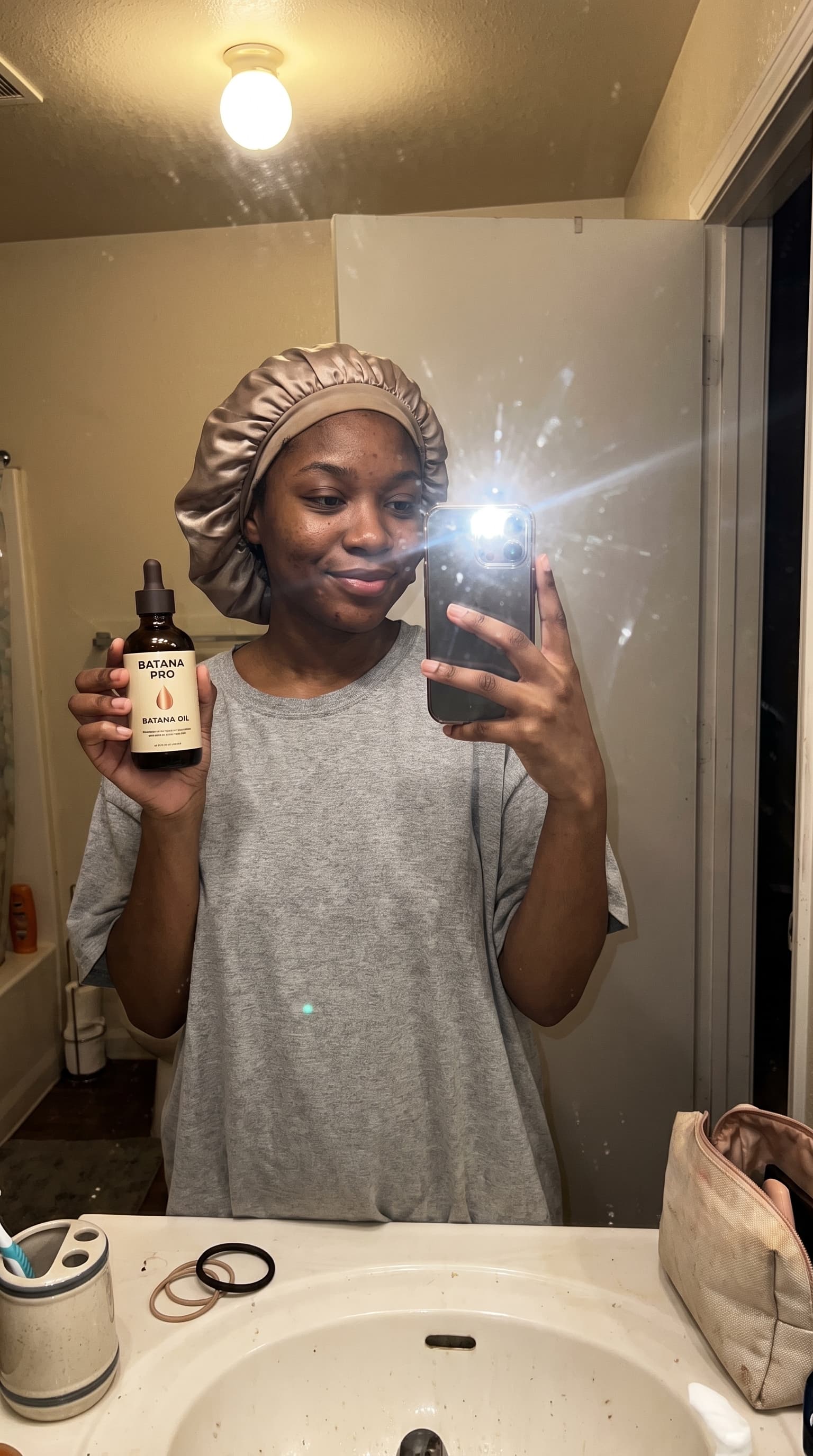 Bathroom mirror selfie at night in a satin bonnet, holding Batana Oil